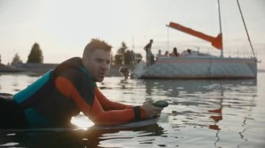 Sörf tahtasında uzanan bir adam marina yakınlarındaki sıcak altın gün batımında dalgıç kıyafeti giyiyor.