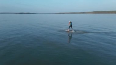 Kamera, büyük mavi gölde, hidrofolyo sörf tahtasının üzerinde bir adamın etrafında uçuyor. 