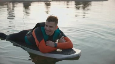 Sıcak altın gün batımında dalgıç kıyafetiyle sörf tahtasına uzanmış gülümseyen bir adam.