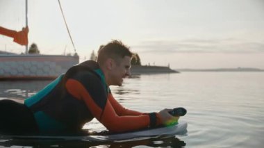 Sörf tahtasında uzanan bir adam marina yakınlarındaki sıcak altın gün batımında dalgıç kıyafeti giyiyor.