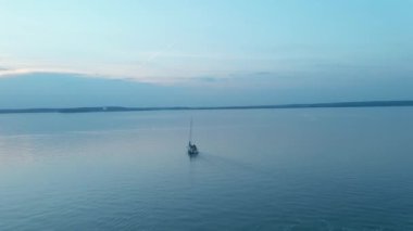 Pembe bir günbatımında mavi bir gölde yelkenlinin üzerinden uçan kamera.