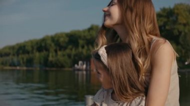 Kızı olan bir kadın gün batımında gülümseyen göle bakarak iskelede oturur.