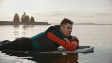 Sıcak altın gün batımında dalgıç kıyafetiyle sörf tahtasına uzanmış gülümseyen bir adam.