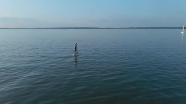 Güneşli bir havada, büyük mavi gölde, su folyosunun üzerinde sörf yapan bir adam.