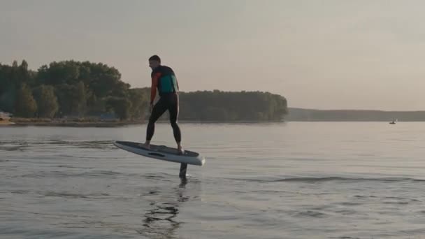 Homem montando em uma prancha de hidrofoil perto da costa do lago e marina ao pôr do sol dourado — Vídeo de Stock