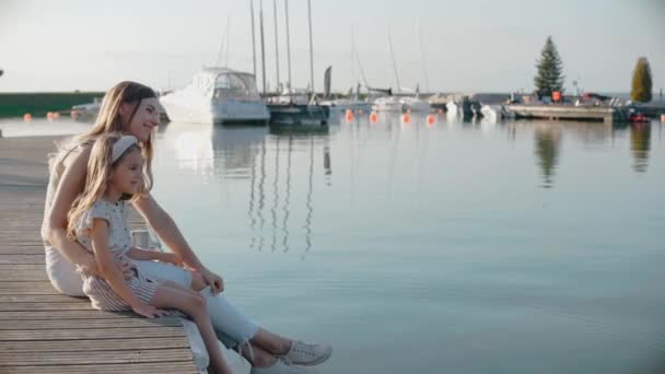 Mulher com filha sentar-se em um cais olhando para o lago ao pôr do sol — Vídeo de Stock