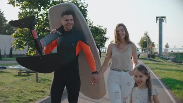 Чоловік гуляє зі своєю сім'єю, несучи дошку для серфінгу на берег озера в сонячний день — стокове відео