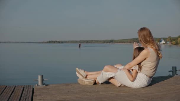 Frau mit Tochter sitzt auf Seebrücke und winkt Papa beim Surfen auf einem See zu — Stockvideo