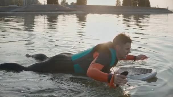 Homem deitado e sorrindo em uma prancha de surf vestindo roupa de mergulho no pôr-do-sol dourado quente — Vídeo de Stock