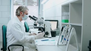 Laboratuvar asistanı mikroskopla çalışıyor ve beyaz laboratuarda notlar yazıyor.