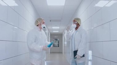 Koruyucu takım elbiseli ve maskeli doktorlar açık beyaz hastane salonunda konuşuyorlar.,