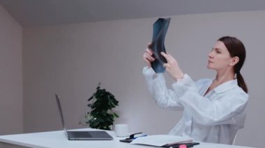 Kadın doktor, parlak ofiste akciğer röntgenlerine bakıyor, üzerinde tıbbi önlük var.