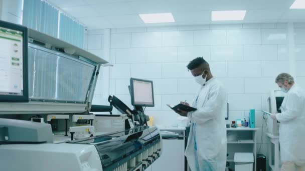 O echipă diversă lucrează în laborator folosind tablete, purtând măști și rochii — Videoclip de stoc