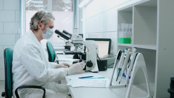 Asistente de laboratorio trabaja con un microscopio y escribe notas en el laboratorio blanco — Vídeos de Stock