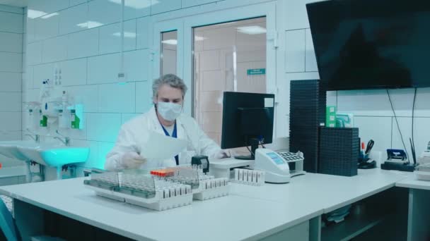 Médecin âgé assis dans un laboratoire avec éprouvettes et travaille sur un ordinateur — Video