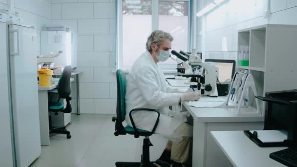 Asistentul de laborator se uită la microscop și scrie note într-un laborator alb — Videoclip de stoc