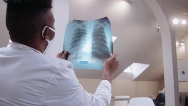 Médecin noir mâle regarde les rayons X pulmonaires dans un bureau lumineux, portant une robe médicale — Video