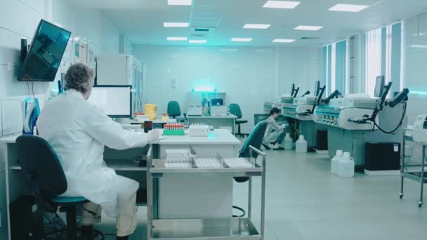 Médico de edad avanzada se sienta en un laboratorio con tubos de ensayo y trabaja en un ordenador — Vídeos de Stock