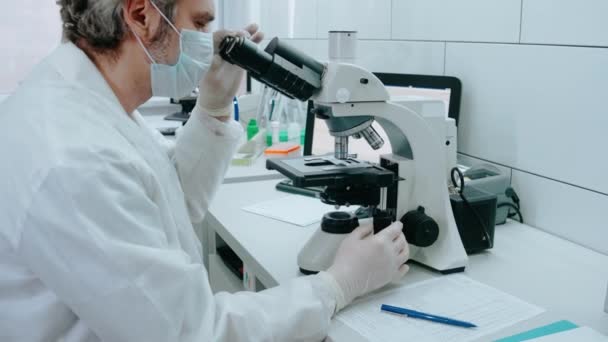 Βοηθός εργαστηρίου κοιτάζει μέσα από ένα μικροσκόπιο και γράφει σημειώσεις σε ένα λευκό εργαστήριο — Αρχείο Βίντεο