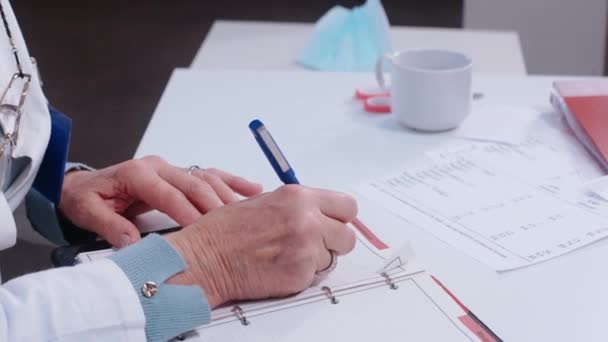 Ηλικιωμένη γυναίκα γιατρός γράφει σημειώσεις κάθεται στο γραφείο, φορώντας ιατρική ρόμπα — Αρχείο Βίντεο