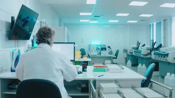 Médico idoso se senta em um laboratório com tubos de ensaio e trabalha em um computador — Vídeo de Stock