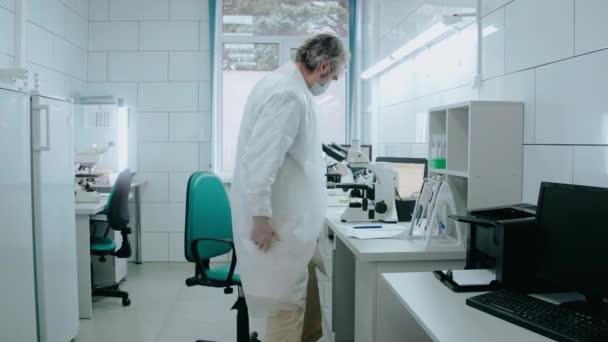 Asistente de laboratorio se sienta en una silla y mira a través de un microscopio en el laboratorio blanco — Vídeos de Stock