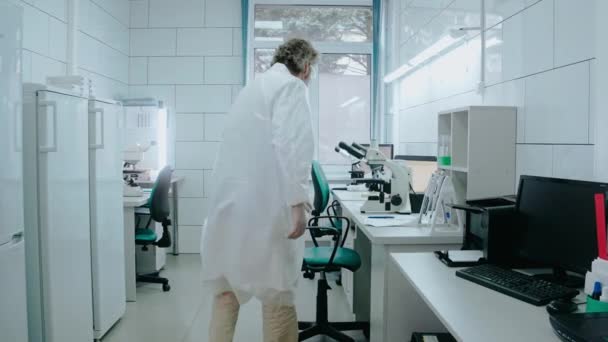 Asystent laboratoryjny siedzi na krześle i pracuje z mikroskopem w białym laboratorium — Wideo stockowe