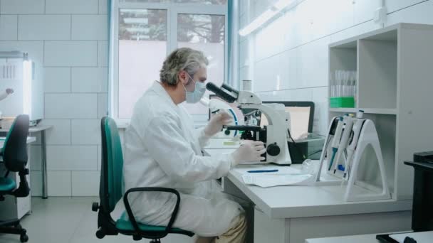 Asistente de laboratorio se sienta en una silla y mira a través de un microscopio en el laboratorio blanco — Vídeos de Stock