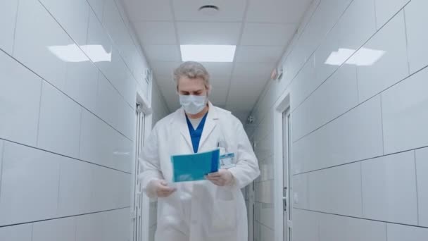 Doctor walks along the white light hospital corridor, reading medical papers — Stock Video