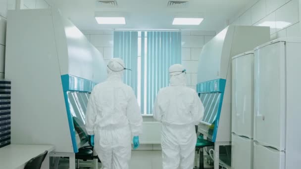 Sanitäter in Schutzanzügen und Masken betreten das Labor und setzen sich auf Stühle — Stockvideo
