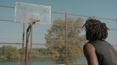 Güneşli bir günde, bir sokak kortunda basketbol topunu sepete atan bir adam.