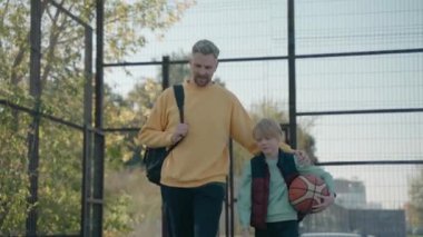 Baba oğul güneşli bir günde basketbol sahasında antreman yapmak için yürüyorlar.