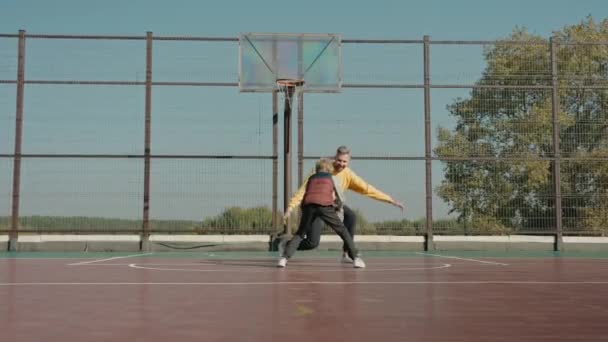 Chłopiec rzucając piłkę gra z ojcem na boisku do koszykówki w słoneczny dzień — Wideo stockowe