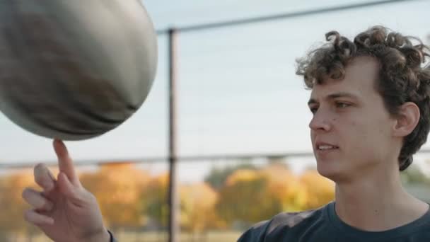 Junge steht auf einem Basketballplatz und dreht an einem sonnigen Tag einen Ball am Finger — Stockvideo
