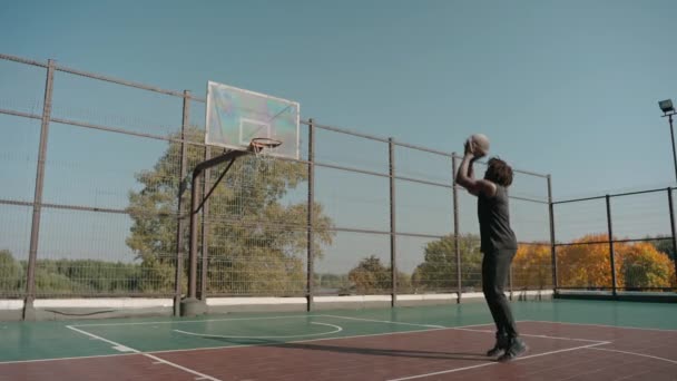 Hombre entrena lanzando pelota de baloncesto en canasta en una cancha callejera en un día soleado — Vídeo de stock