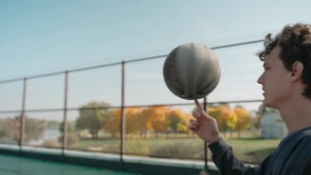 Boy stands on a basketball court and spins a ball on his finger on a sunny day — Stock Video