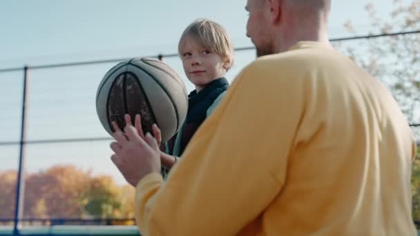 Vater erklärt Sohn, wie man an einem sonnigen Tag einen Basketballball hält und wirft — Stockvideo