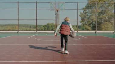 Sahada antrenman yapan bir çocuk parkta basketbol topu atıyor.