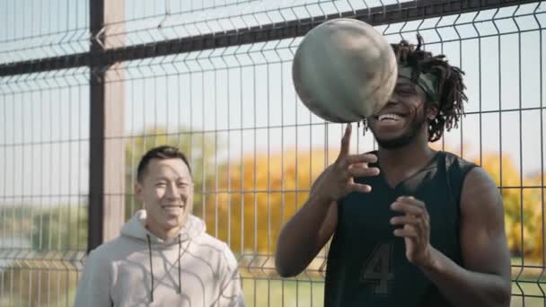 Czarny mężczyzna kręci piłką na palcu, a jego przyjaciel uśmiecha się na boisku. — Wideo stockowe