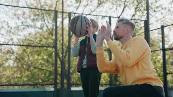 Vater erklärt Sohn, wie man an einem sonnigen Tag einen Basketballball hält und wirft — Stockvideo