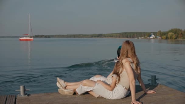 Γυναίκα με κόρη κάθονται σε μια προβλήτα και βλέπουν τον μπαμπά να σερφάρει σε μια λίμνη στο ηλιοβασίλεμα. — Αρχείο Βίντεο