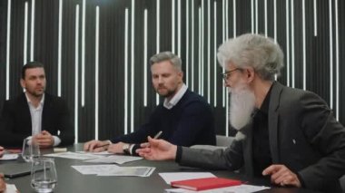 Takım elbiseli yaşlı işadamı toplantıda oturan meslektaşları veya ortaklarıyla konuşuyor