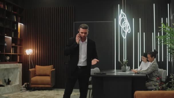 Чоловік у костюмі сперечається по телефону в темному офісі, співробітники сидять за столом — стокове відео