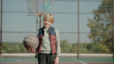 Çocuk elinde basketbol topuyla sahada duruyor ve kameraya bakıyor.