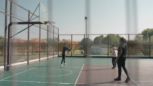 Trzech młodych mężczyzn rzuca piłkę do kosza na boisku w parku w słoneczny dzień — Wideo stockowe