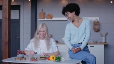 Genç kız masada oturan kız arkadaşıyla sebze doğruyor ve gülüyor.