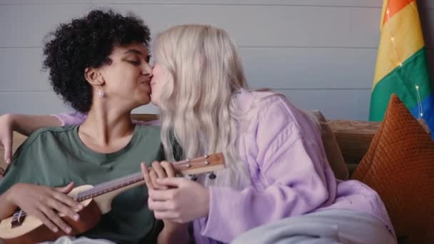 Las chicas jóvenes se besan sentadas en un sofá y tocando el ukelele — Vídeos de Stock
