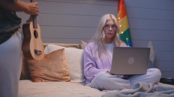 Dívka přijde ke své přítelkyni sedí v posteli, pracuje na notebooku hrát ukulele — Stock video