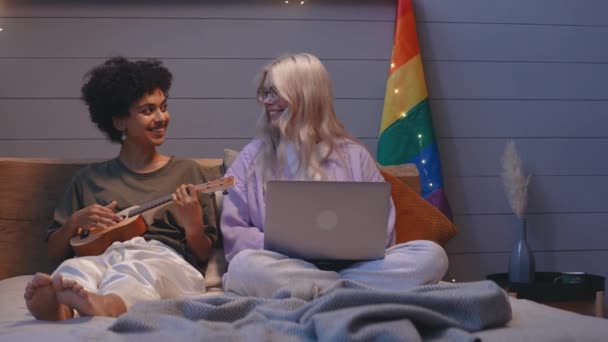 Joven pareja lgbt sentarse en la cama, trabajando en un ordenador portátil y jugando en ukelele — Vídeos de Stock
