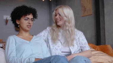 Lezbiyen çift konuşuyor, gülüyor, birbirlerine bakıyorlar, evdeki kanepede oturuyorlar.